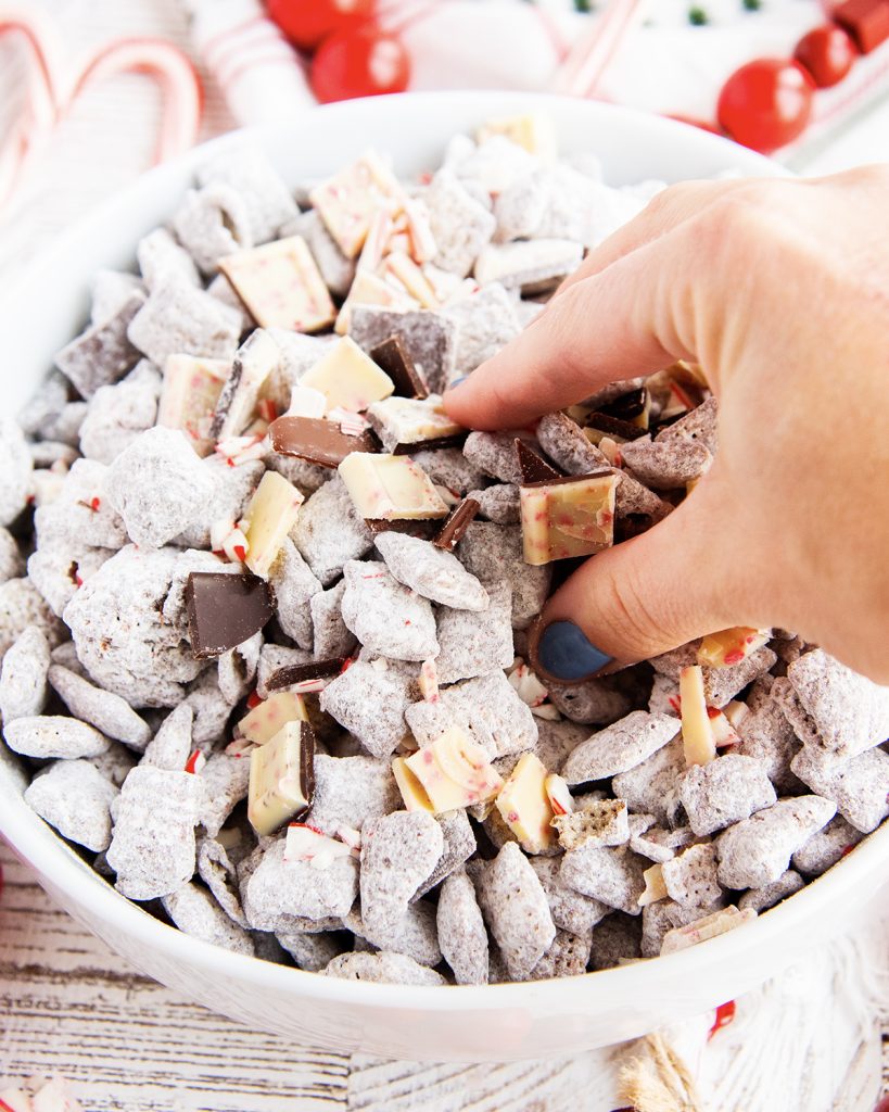 A hand grabbing into a bowl of peppermint bark muddy buddies.
