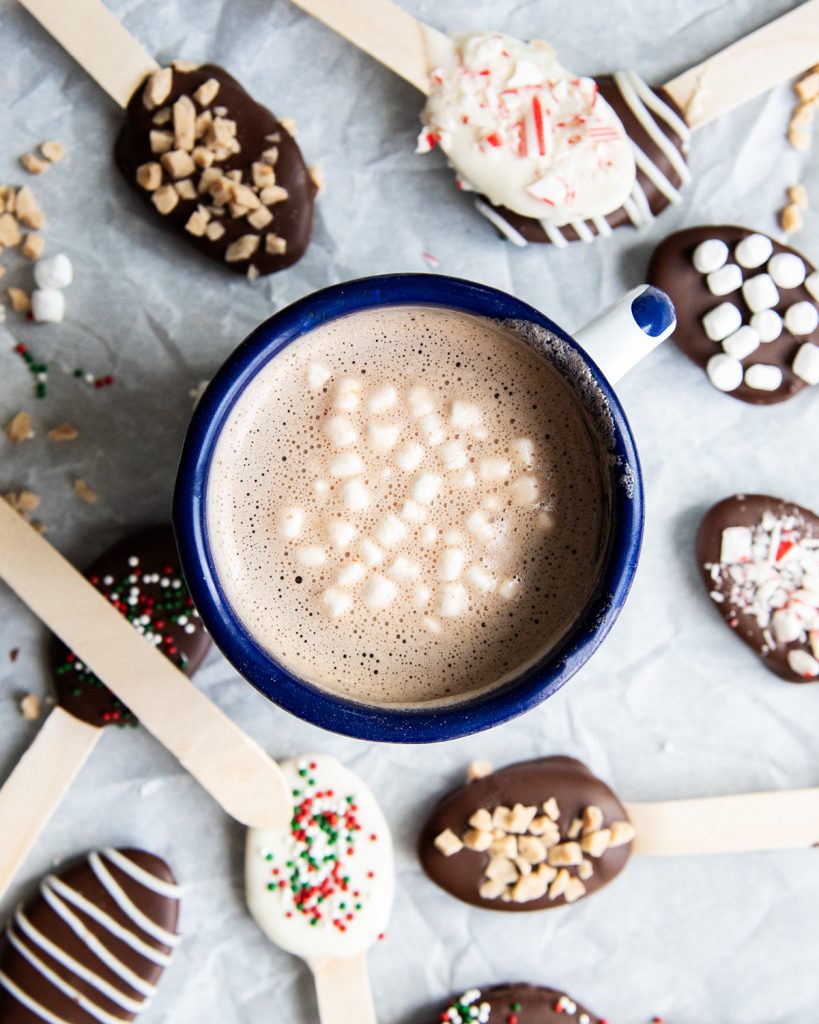 A cup of hot cocoa topped with mini marshmallows. With hot chocolate spoons around the mug.