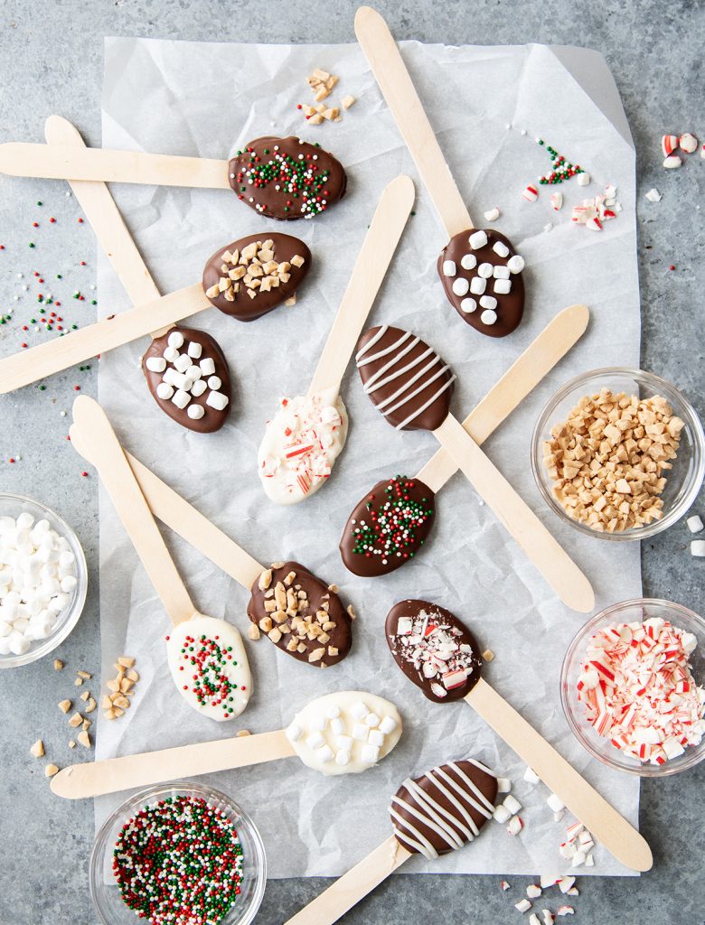An overhead photo of hot chocolate spoons, with the toppings for them in small bowls around them, sprinkles, toffee bits, candy cane pieces, and mini marshmallows.