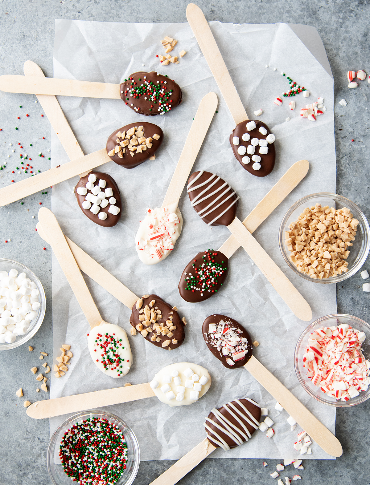 An overhead photo of hot chocolate spoons, with the toppings for them in small bowls around them, sprinkles, toffee bits, candy cane pieces, and mini marshmallows.