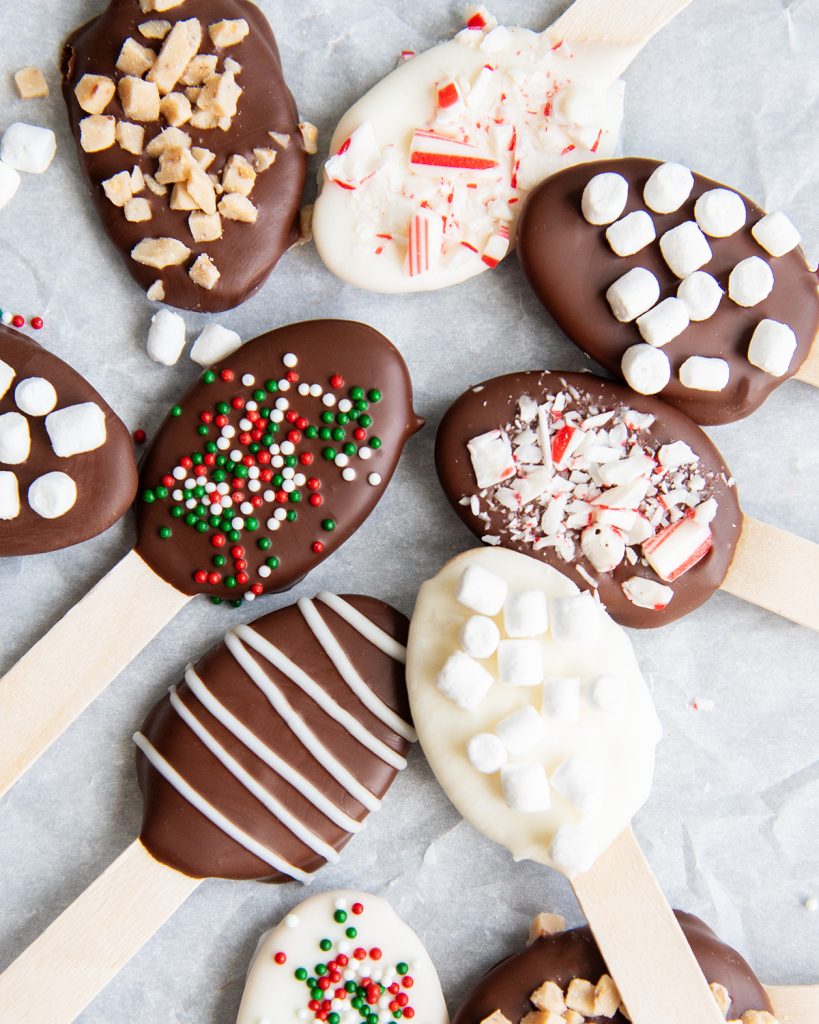 Hot chocolate spoons arranged in a random pile, some have sprinkles on them, some have mini marshmallows, some have candy cane pieces.