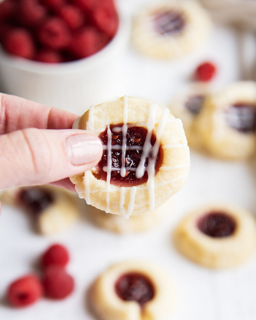 A hand holding a raspberry thumbprint cookie, drizzled with an almond icing.