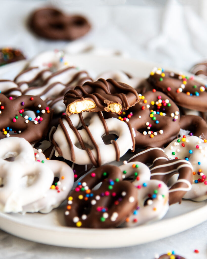 A plate of chocolate covered pretzels, and one has a bite out of it, showing the pretzel in the middle.
