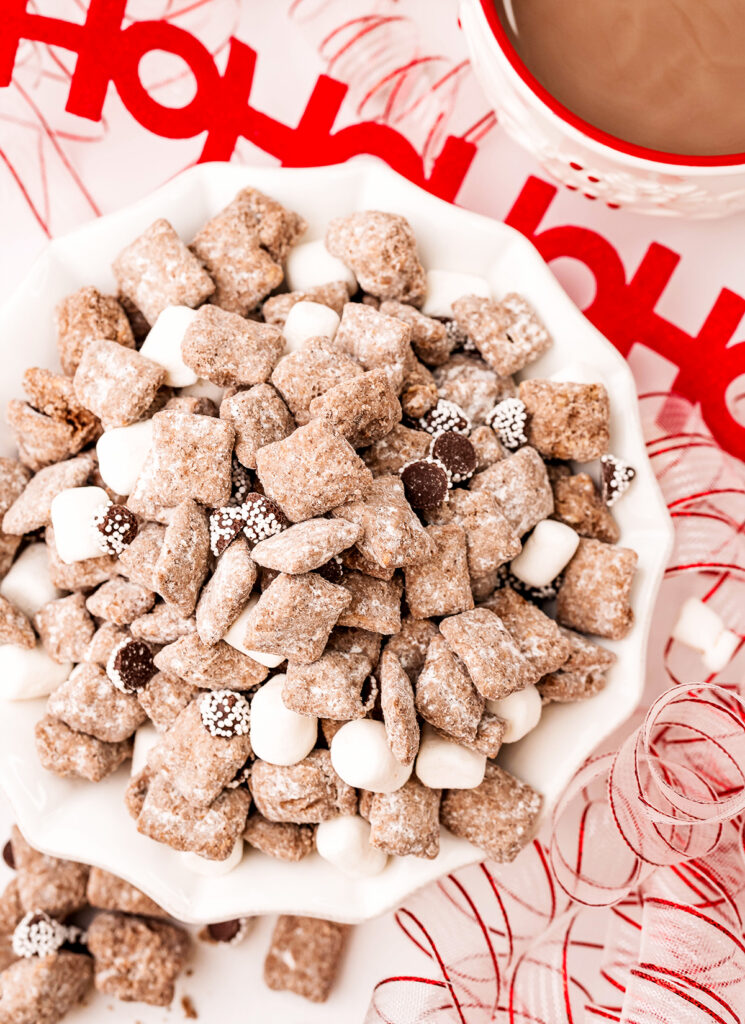 Pieces of chocolate covered muddy buddies, Snow Cap candies, and mini marshmallows on a surface, with a bowl of the mixture behind.