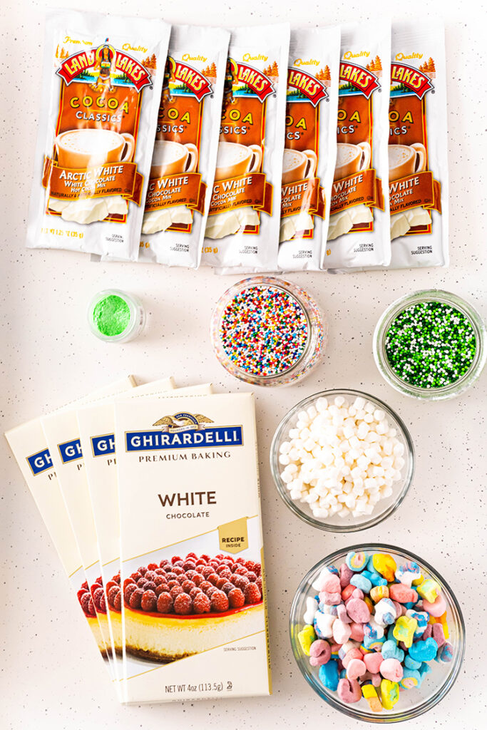The ingredients needed to make white hot chocolate bombs. There are packs of white chocolate cocoa mix, white chocolate baking chocolate bar, bowls or sprinkes, a bowl of mini marshmallows, and a bowl of lucky charms marshmallows.