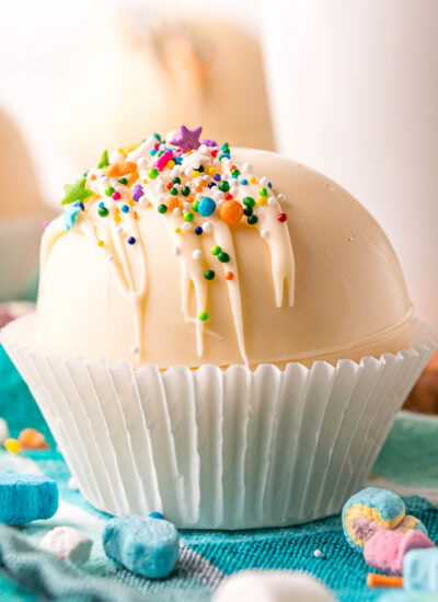 A close up side view of a white chocolate cocoa bomb in a white muffin liner, topped with colorful sprinkles.