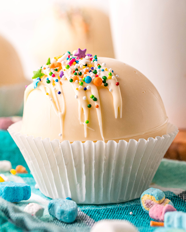 A close up side view of a white chocolate cocoa bomb in a white muffin liner, topped with colorful sprinkles. 