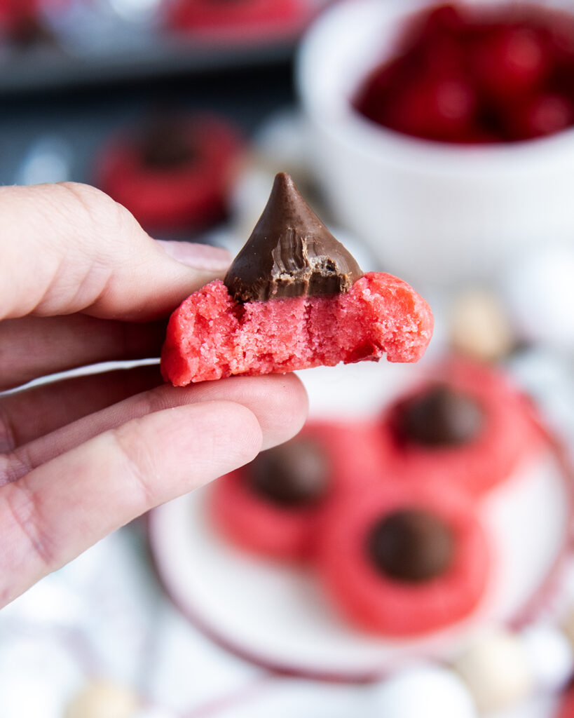 A hand holding a bite sized cherry kiss cookie, with a bite out of them.