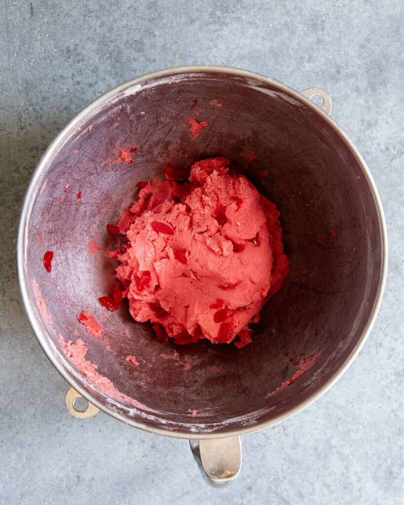 A mixing bowl full of red cookie dough, with chunks of Maraschino cherries.