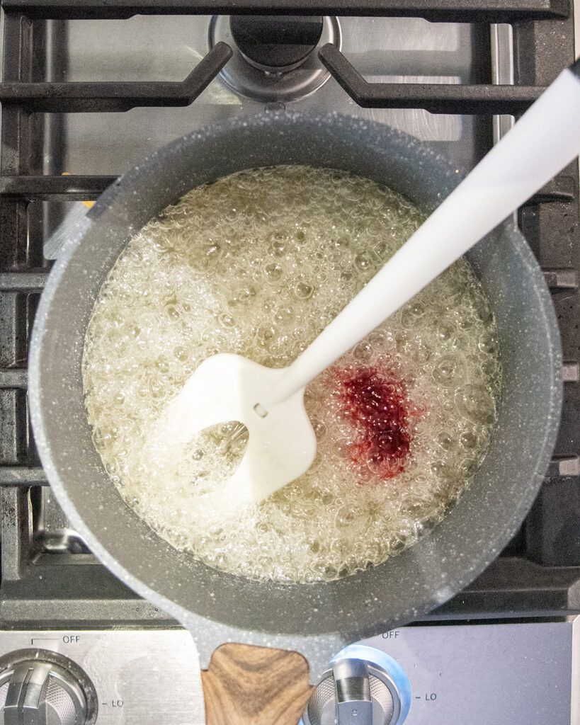 A boiling sugar mixture in a pot with a drop of red food coloring in it.