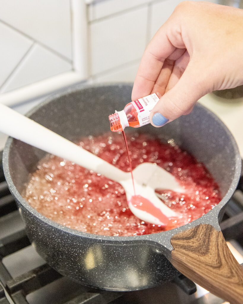 A hand pouring a bottle of flavoring into a pot of pink liquid.