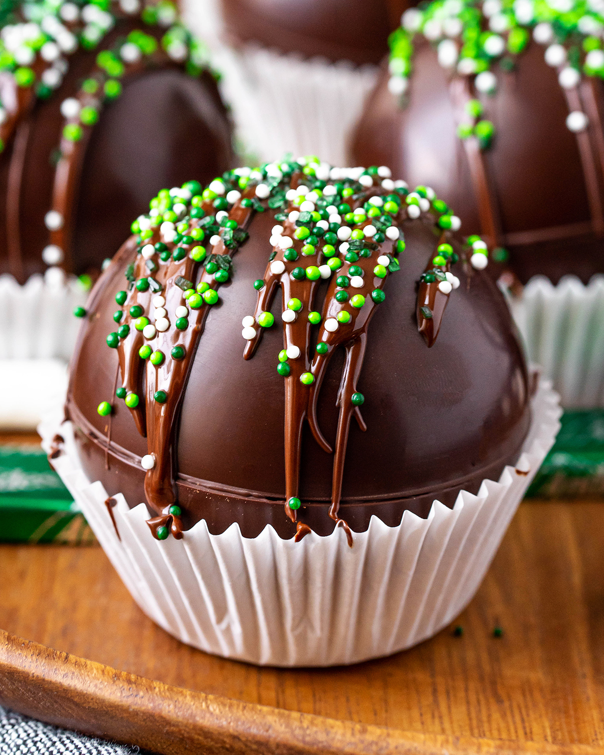 A chocolate hot cocoa bomb topped with a drizzle of chocolate and green and white sprinkles.