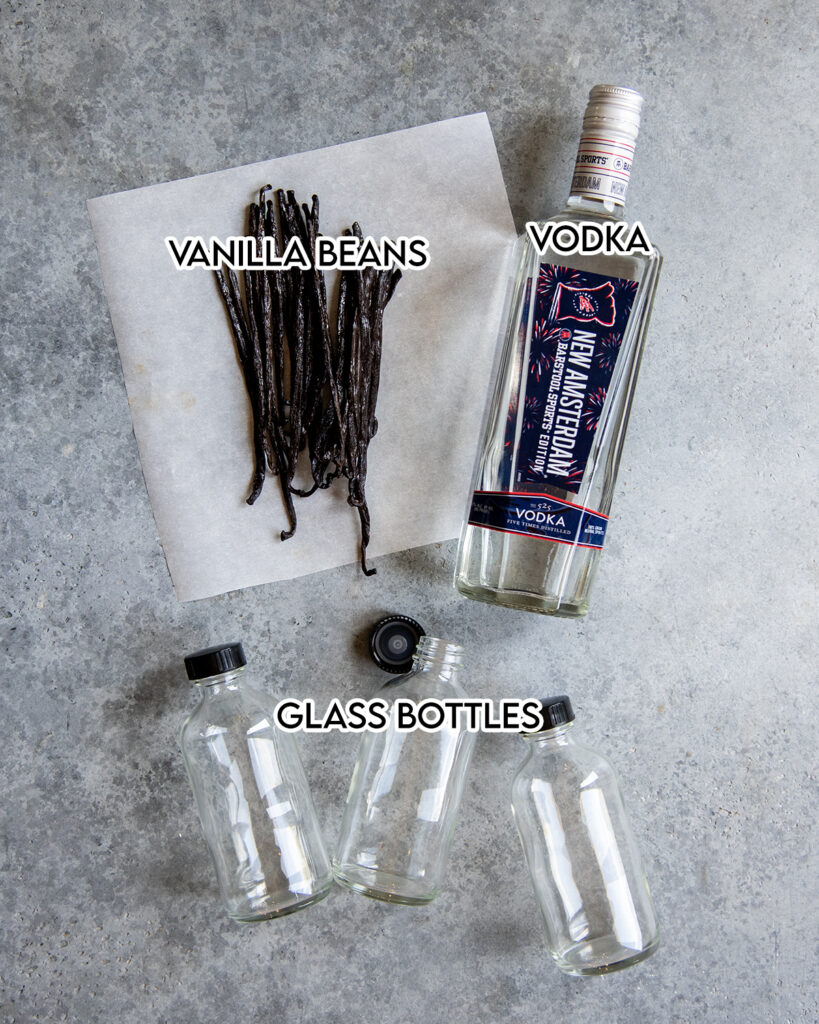 An above photo of a bottle of vodka, vanilla beans on a piece of parchment paper, and three glass bottles. 