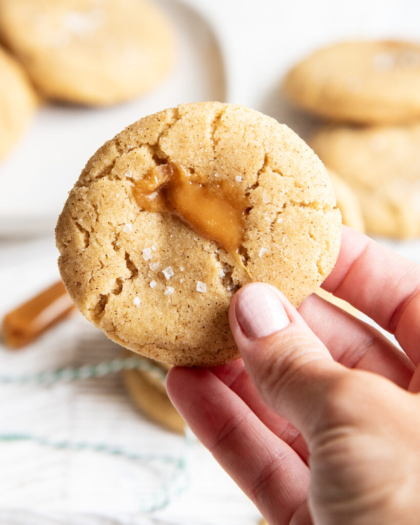 A hand holding a snickerdoodle cookie with caramel seeping out the top. 