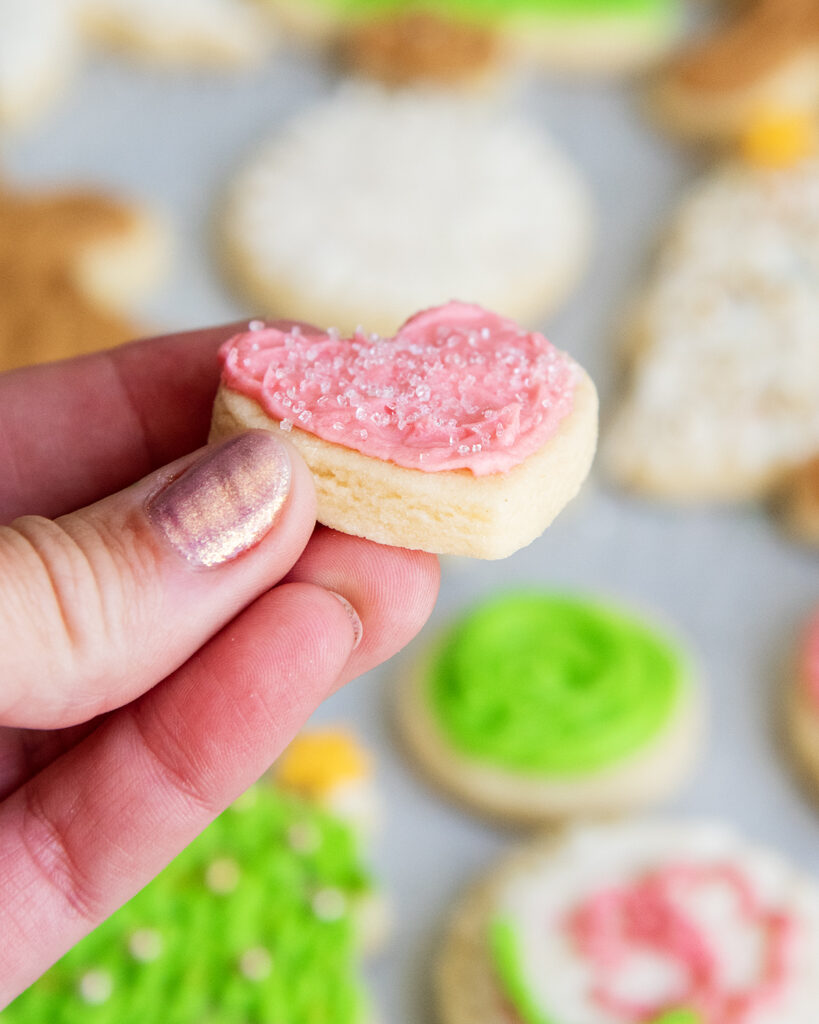 A hand holding a tiny heart shaped sugar cookie with pink frosting.