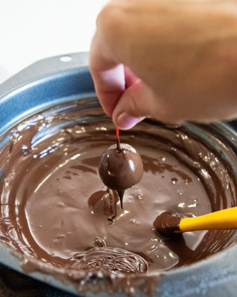 A hand holding a cherry dipped in chocolate and lifting it out of a bowl of melted chocolate. 