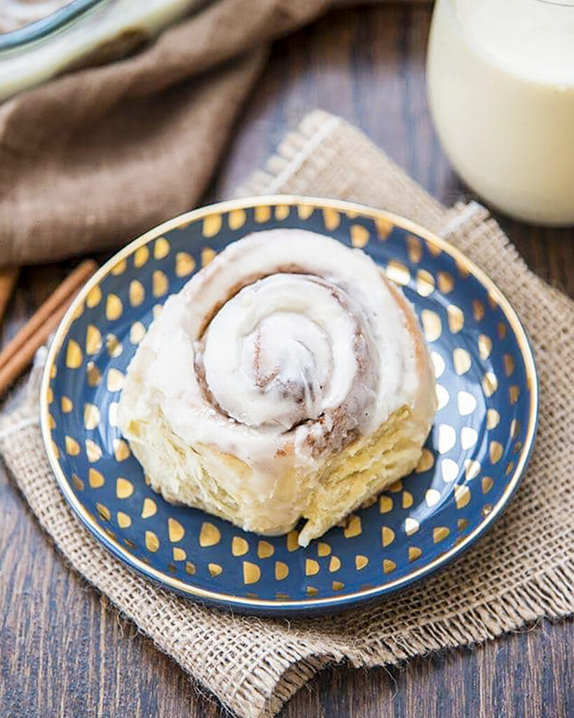 A blue plate topped with an eggnog cinnamon roll, next to a cup of eggnog.