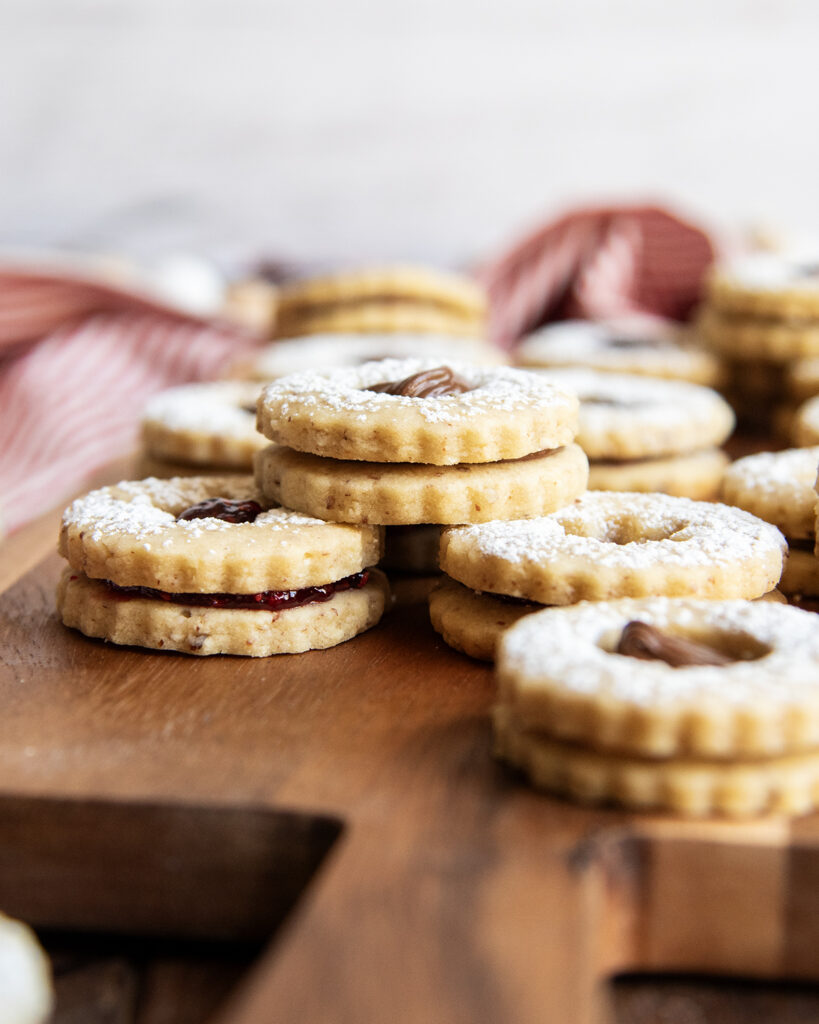 A pile of sandwich cookies filled with Nutella and jam on a wooden board.