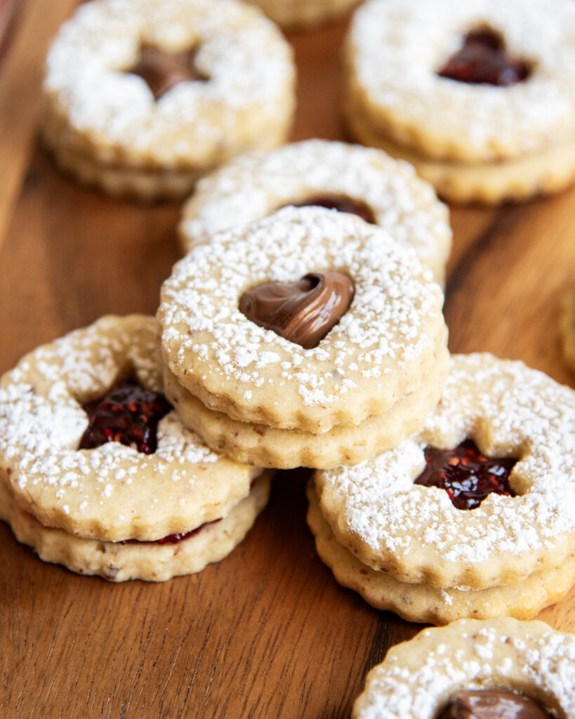 A pile of sandwich cookies with cut outs on the top cookie, they are filled with jam and chocolate hazelnut spread. 