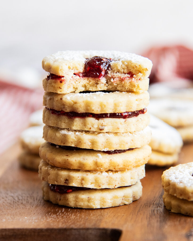 A stack of sandwich cookies and the top cookie has a bite out of it showing raspberry jam in the middle.