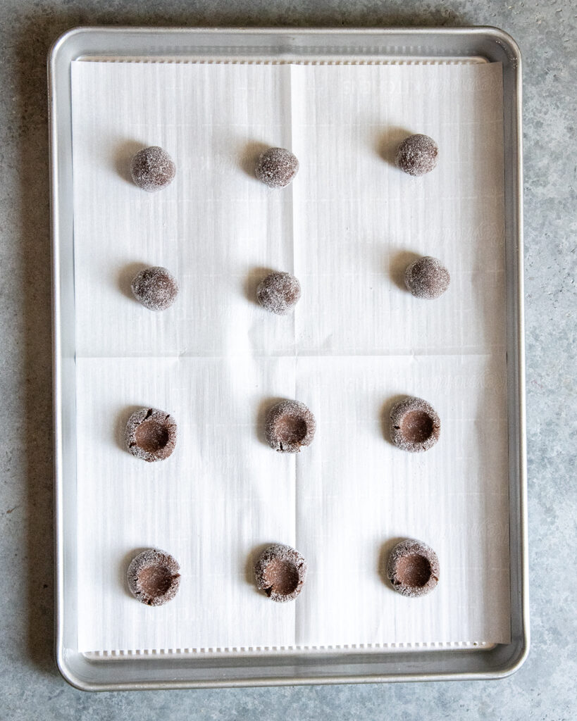 A cookie sheet with balls of chocolate cookie dough, and half of them have thumbprints in the middle.