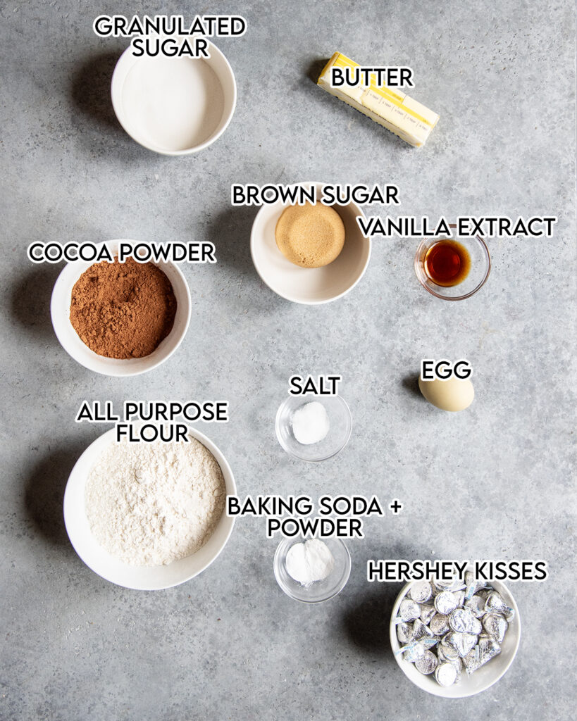 Bowls of the ingredients needed to make chocolate blossom cookies all in small bowls. There is granulated sugar, brown sugar, cocoa powder, flour, an egg, hershey's kisses, and more. 