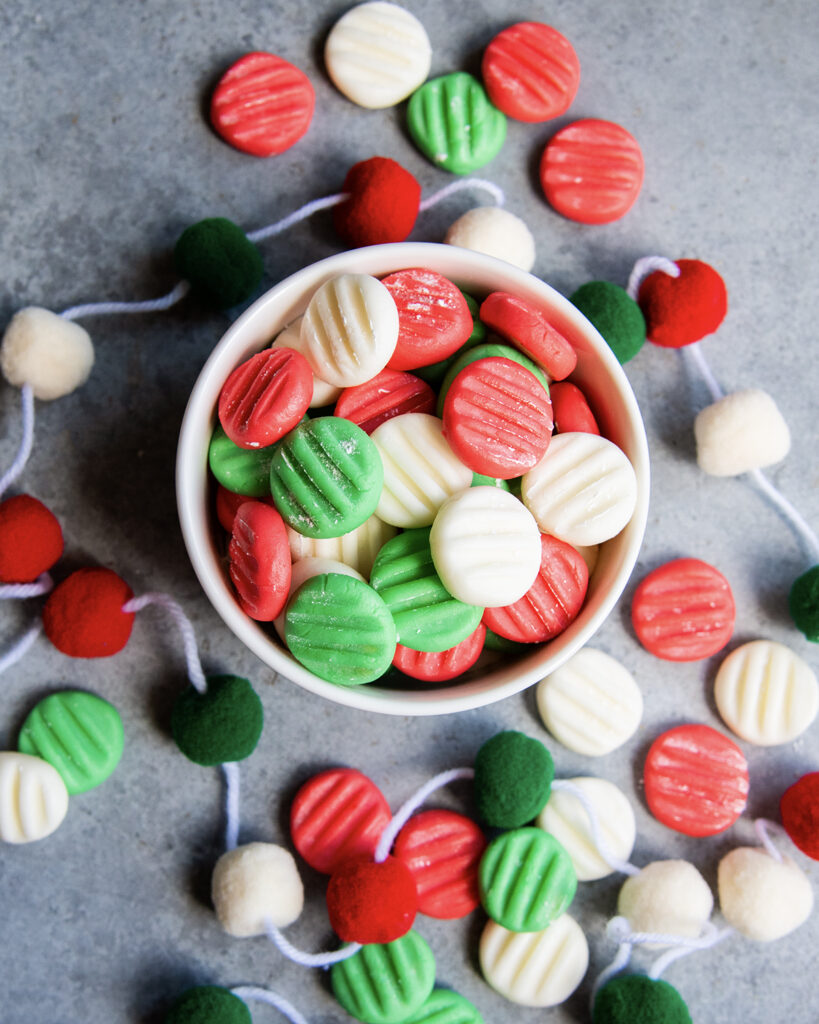 A bowl of red, white, and green cream cheese mints with some around the bowl too.