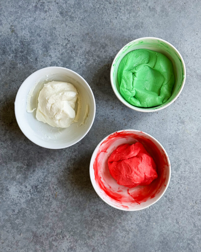 Three bowls of a powdered sugar cream cheese mixture, one is green, one is red, and one is white.