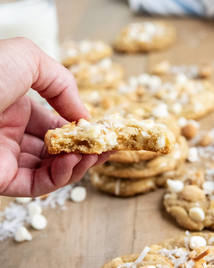 A hand holding half a white chocolate chip cookie, with bites out of it.