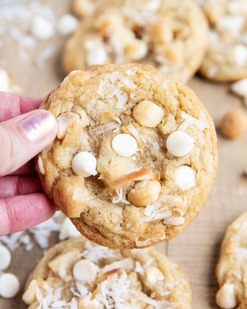 A hand holding a white chocolate chip, coconut, and macadamia nut cookie.