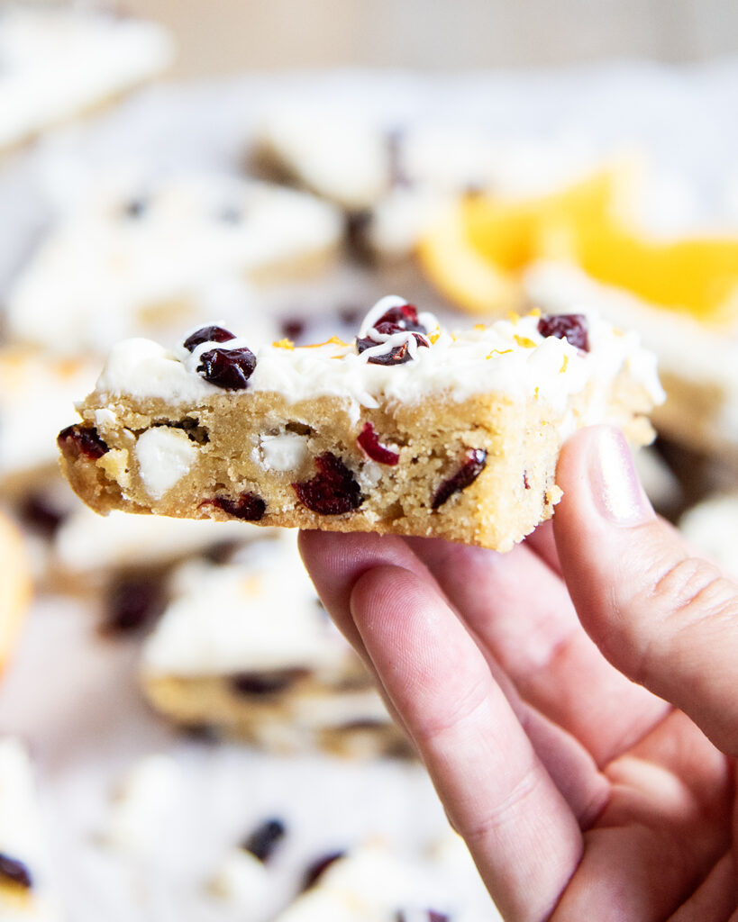 A hand holding a homemade cranberry bliss bar, with white chocolate and cranberry pieces, and a cream cheese frosting.