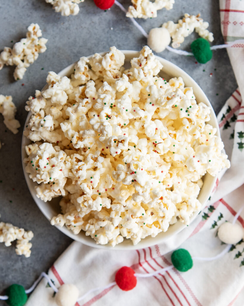 An above view of a bowl of white chocolate coated popcorn, topped with tiny round sprinkles. 