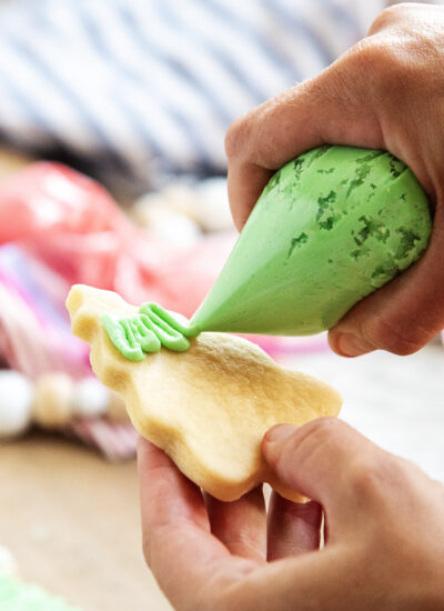 A hand piping green frosting onto a tree shaped sugar cookie.