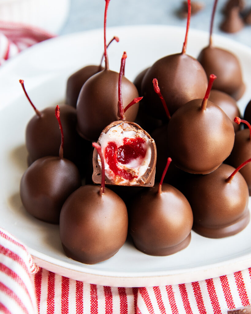 A plate of chocolate covered cherries in a pile, and one has a bite out of it, showing the maraschino cherry in the middle.