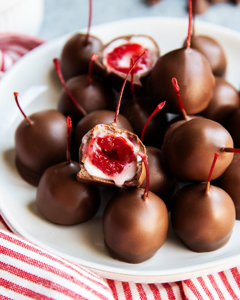 A plate of chocolate covered cherries in a pile, and two have a bite out of them, showing the maraschino cherry in the middle.