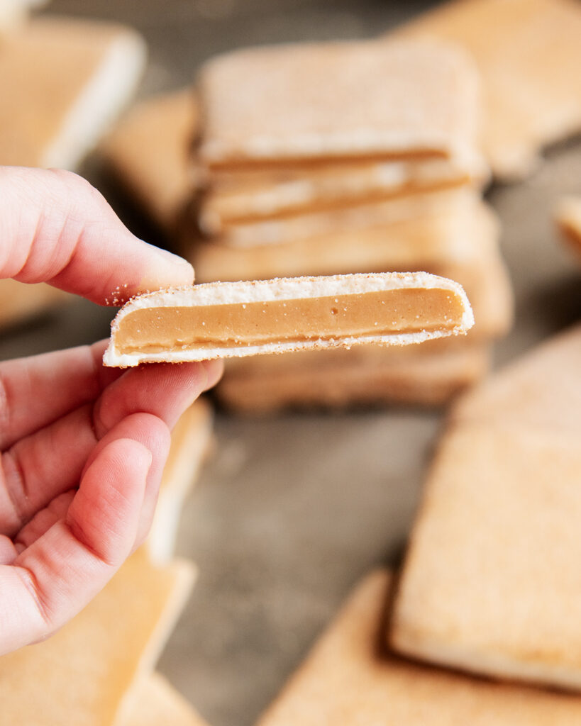 A hand holding a piece of churro toffee, showing the golden toffee in the middle coated with white chocolate.