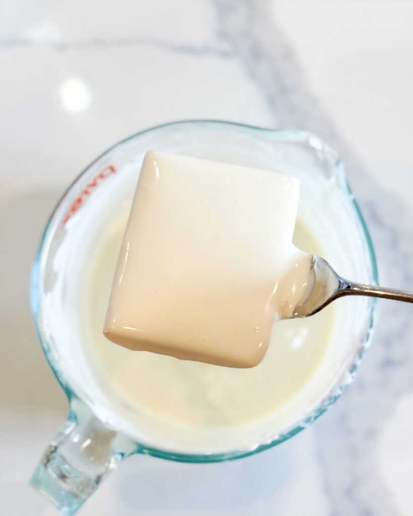 A square of toffee dipped in melted white chocolate above a bowl of it.