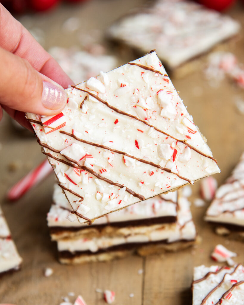 A hand holding a piece of peppermint cracker toffee, it looks like white chocolate peppermint bark with candy cane pieces on top.