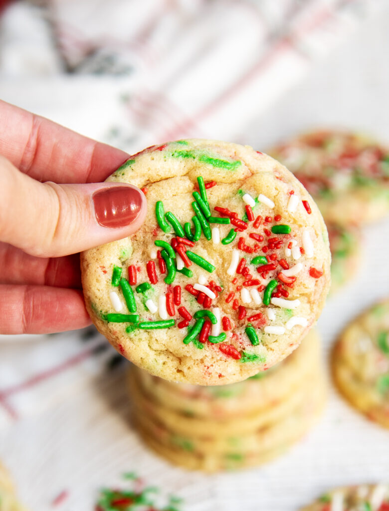 A hand holding a Christmas sprinkle sugar cookie topped with red, white, and green jimmie sprinkles.