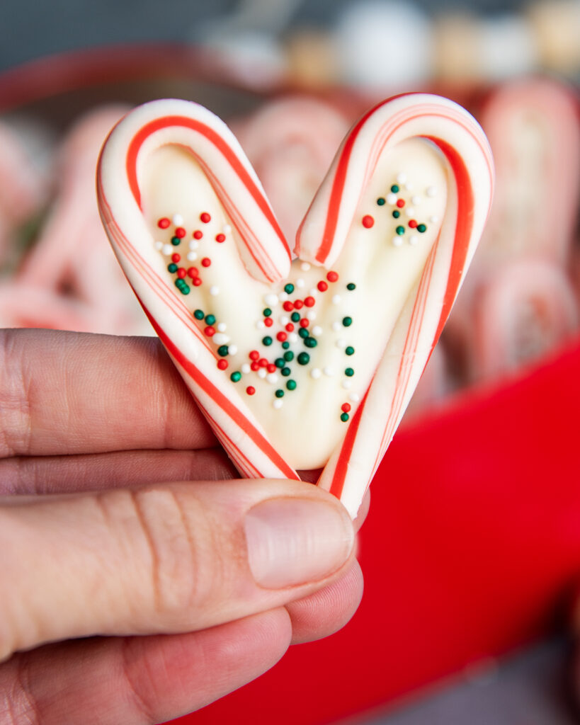 A hand holding a candy cane heart filled with white chocolate and topped with holiday sprinkles.