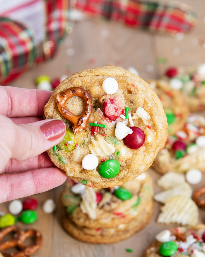 A hand holding a Kitchen Sink Cookie, topped with white chocolate chips, Christmas m&ms, and pretzels.