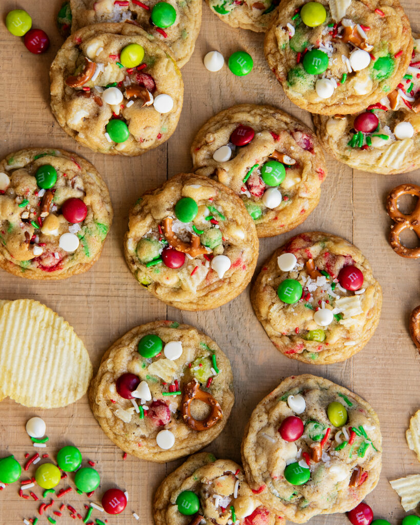 A pile of Christmas Kitchen Sink Cookies on a wooden table. They are topped with Christmas m&ms, white chocolate chips, pretzels, and ruffles chips.