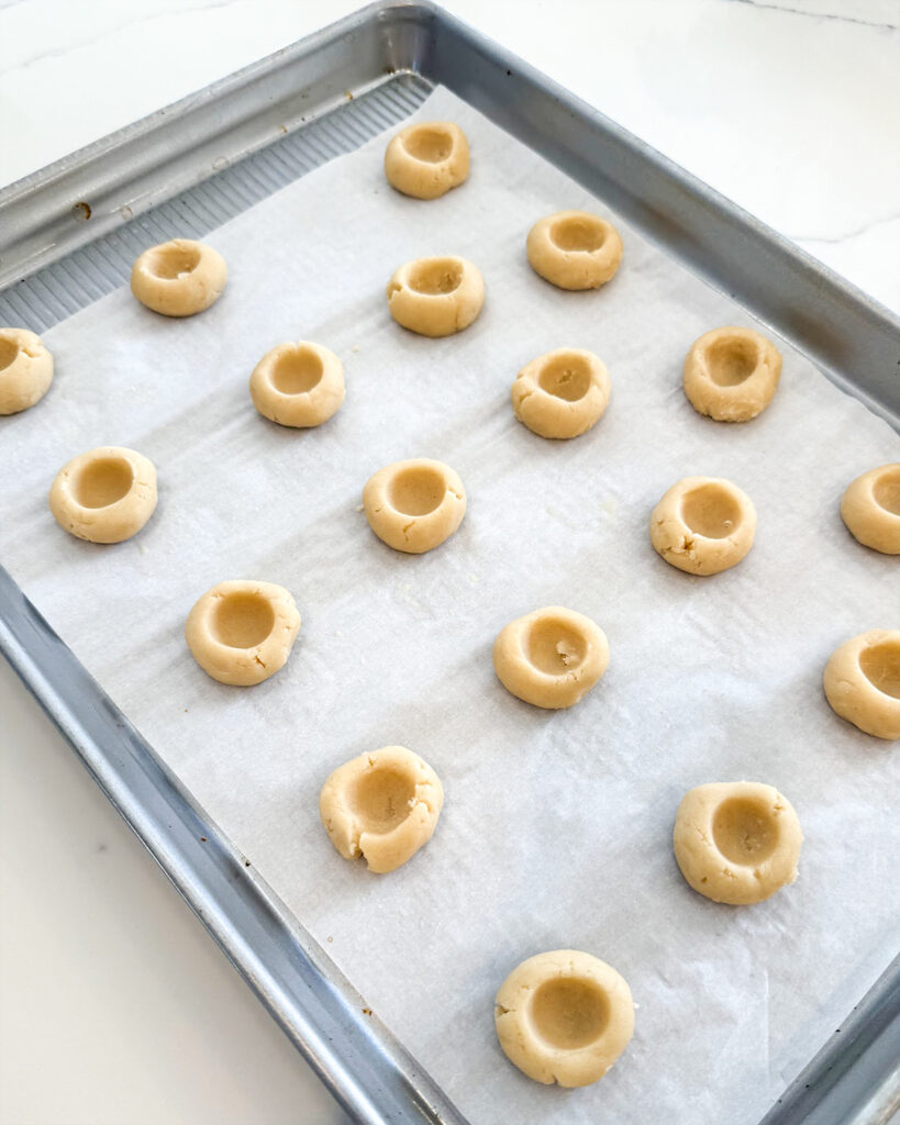 Thumbprint cookie dough on a cookie sheet, with the thumb indents in the dough balls.