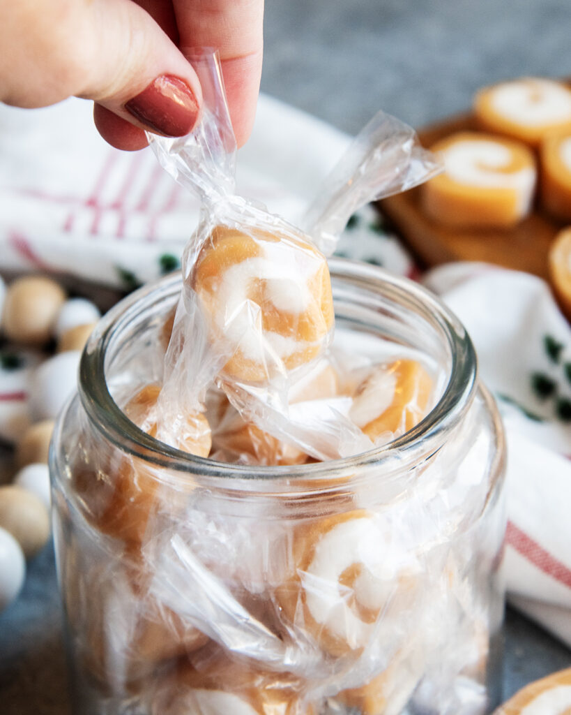 A hand lifting a candy out of a jar of packaged vanilla caramel creams.