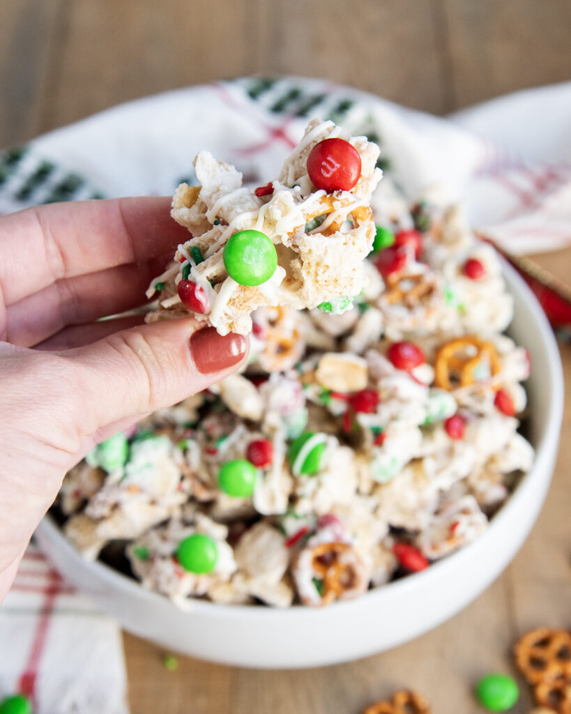 A hand holding a piece of white chocolate coated chex mix topped with peanuts and m&ms.