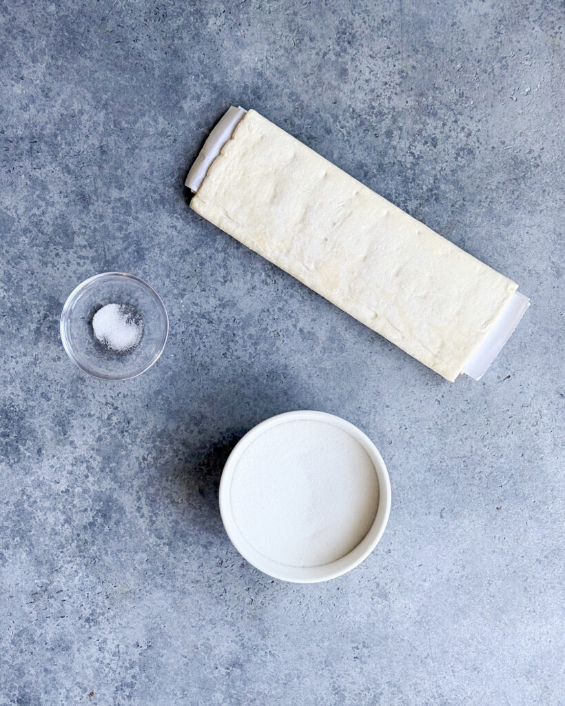 Puff Pastry, a bowl of salt, and a bowl of sugar on a gray counter top. 