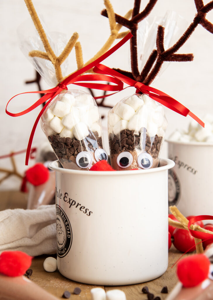 Two Reindeer Hot Chocolate packs in reindeer decorated bags sitting in a porcelain mug.