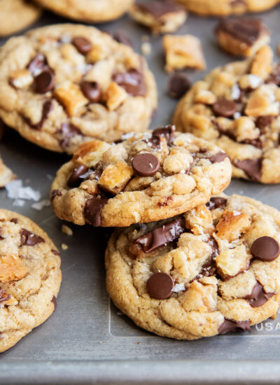 An above view of chocolate chip cookies on a cookie sheet with pieces of cracker toffee in them, and topped with flaky sea salt.