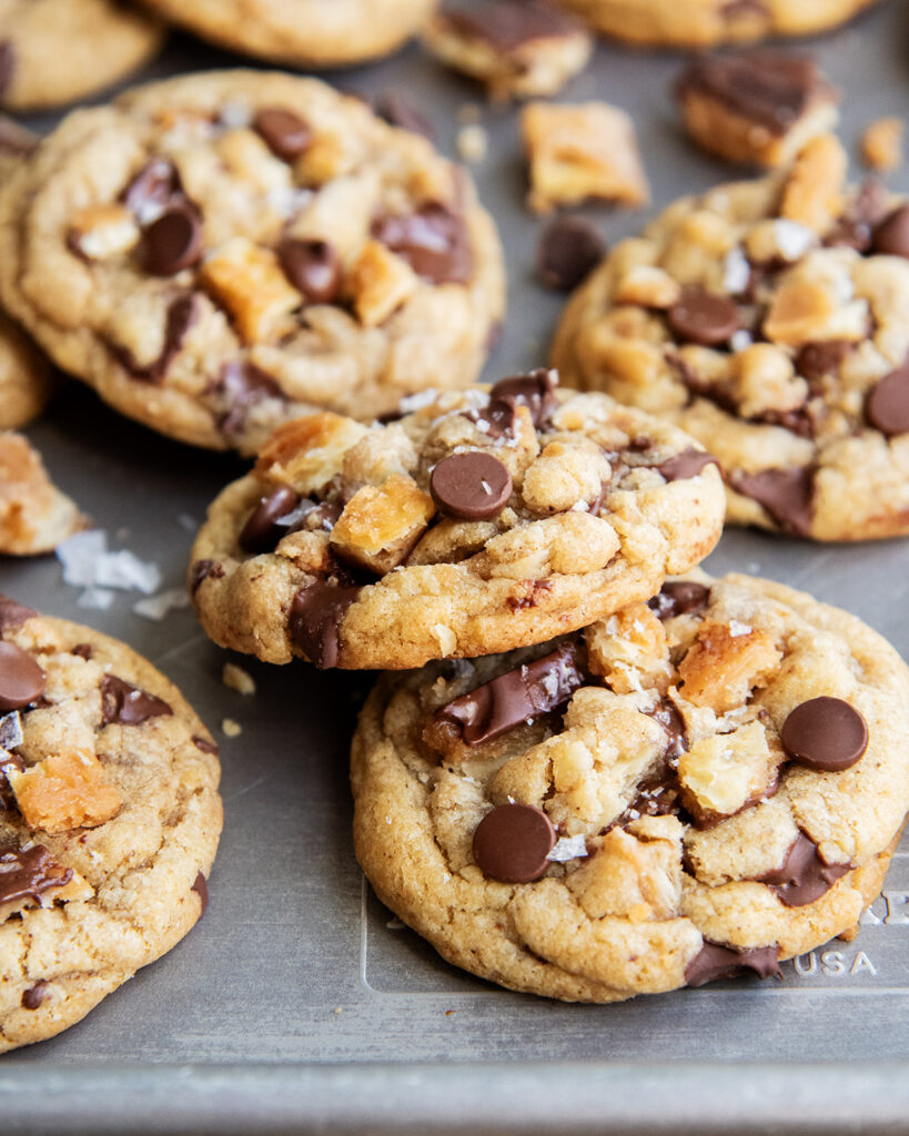 An above view of chocolate chip cookies on a cookie sheet with pieces of cracker toffee in them, and topped with flaky sea salt.