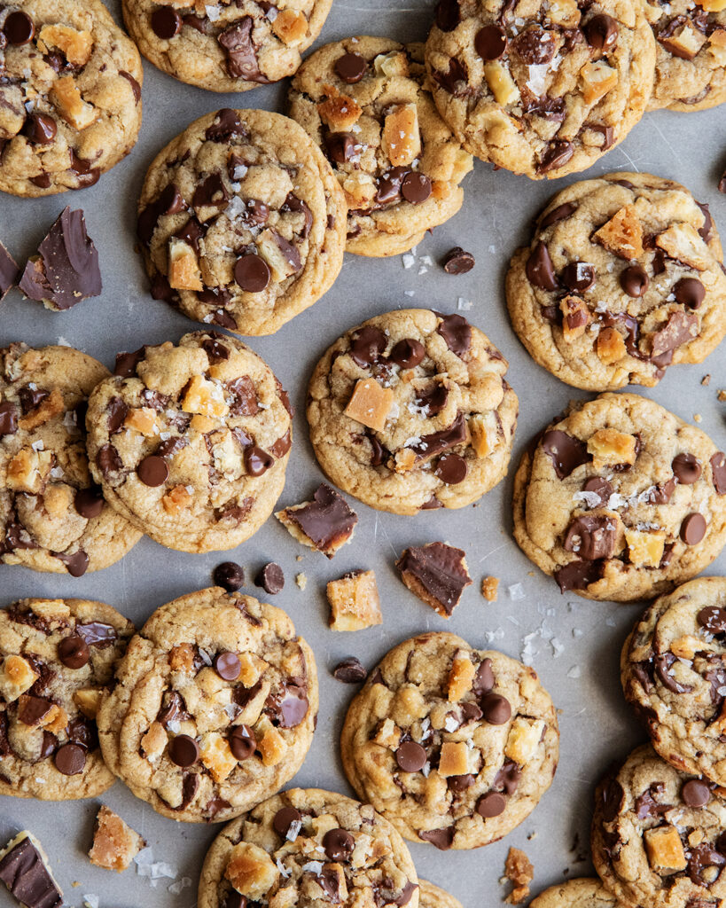 An above view of chocolate chip cookies on a cookie sheet with pieces of cracker toffee in them, and topped with flaky sea salt.